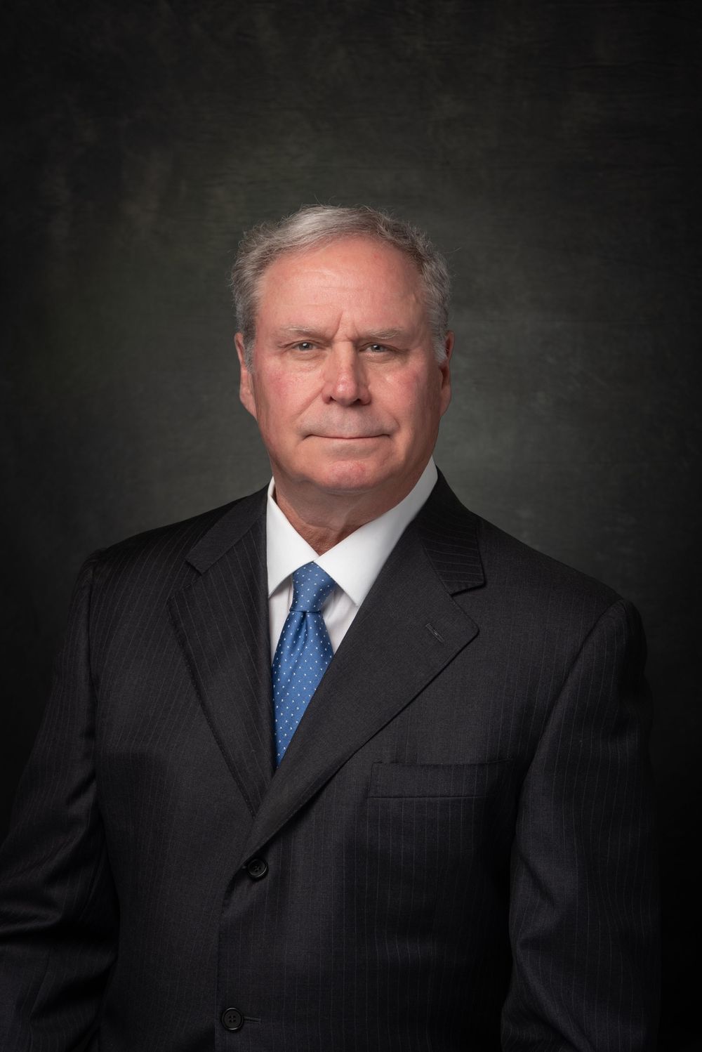 Business headshot of older man with gray hair, in a  black suit with blue tie and white shirt.