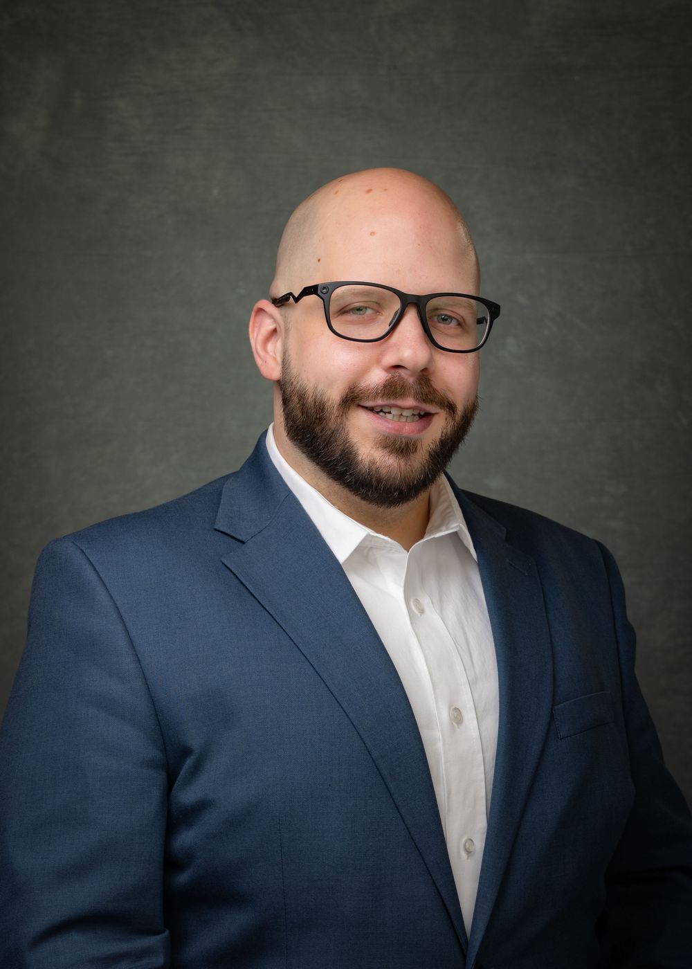 Business Headshot of man in a blue suit and white shirt with glasses and a beard.