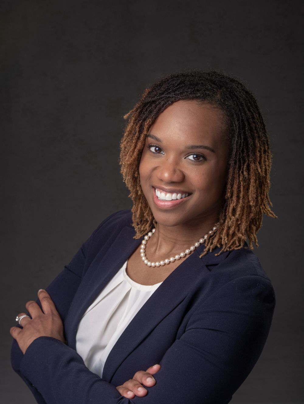 Professional headshot of black woman in a navy business suit and white top with arms crossed.