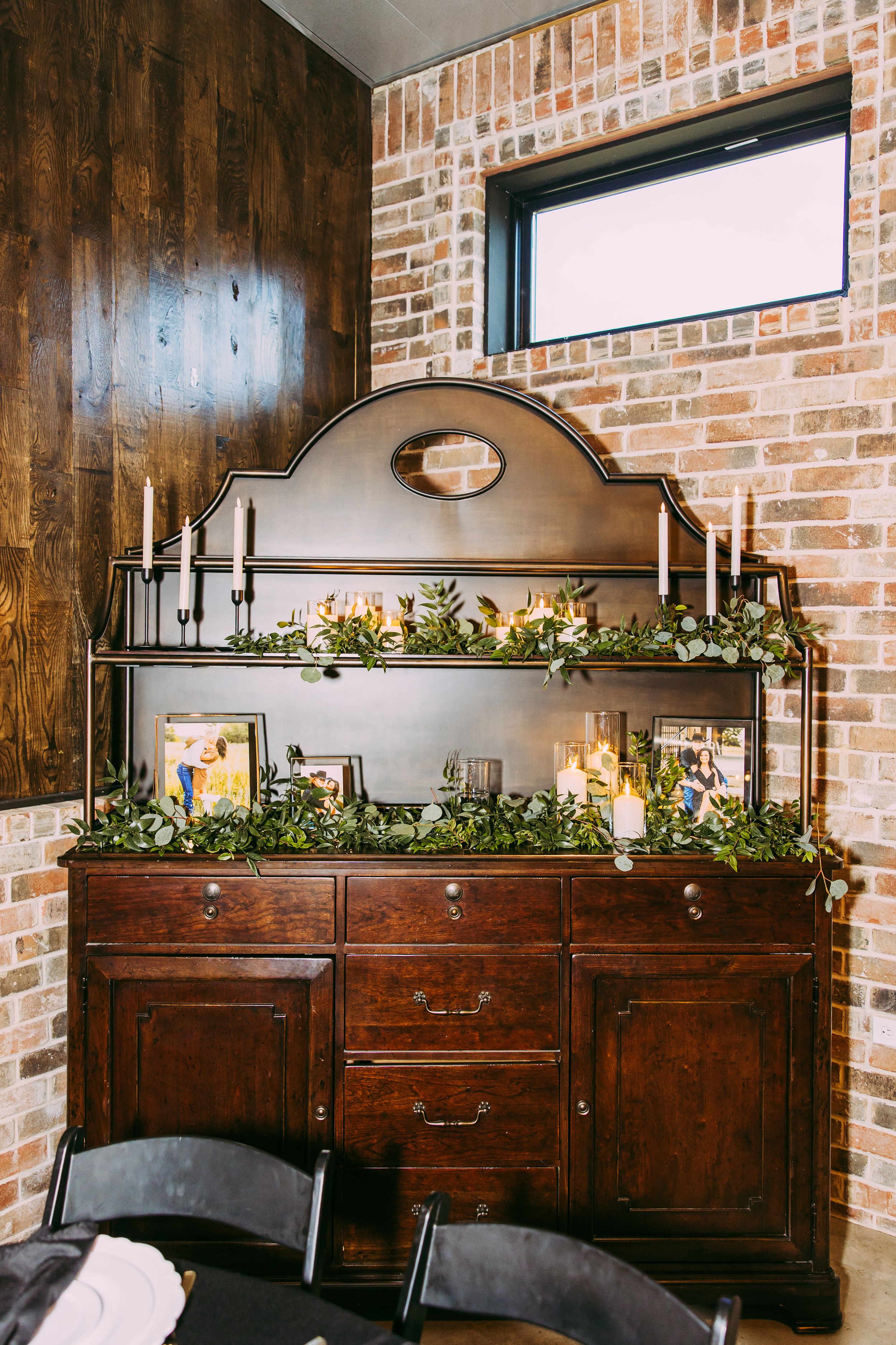 Vintage wooden buffet decorated with greenery and candles against exposed brick wall.
