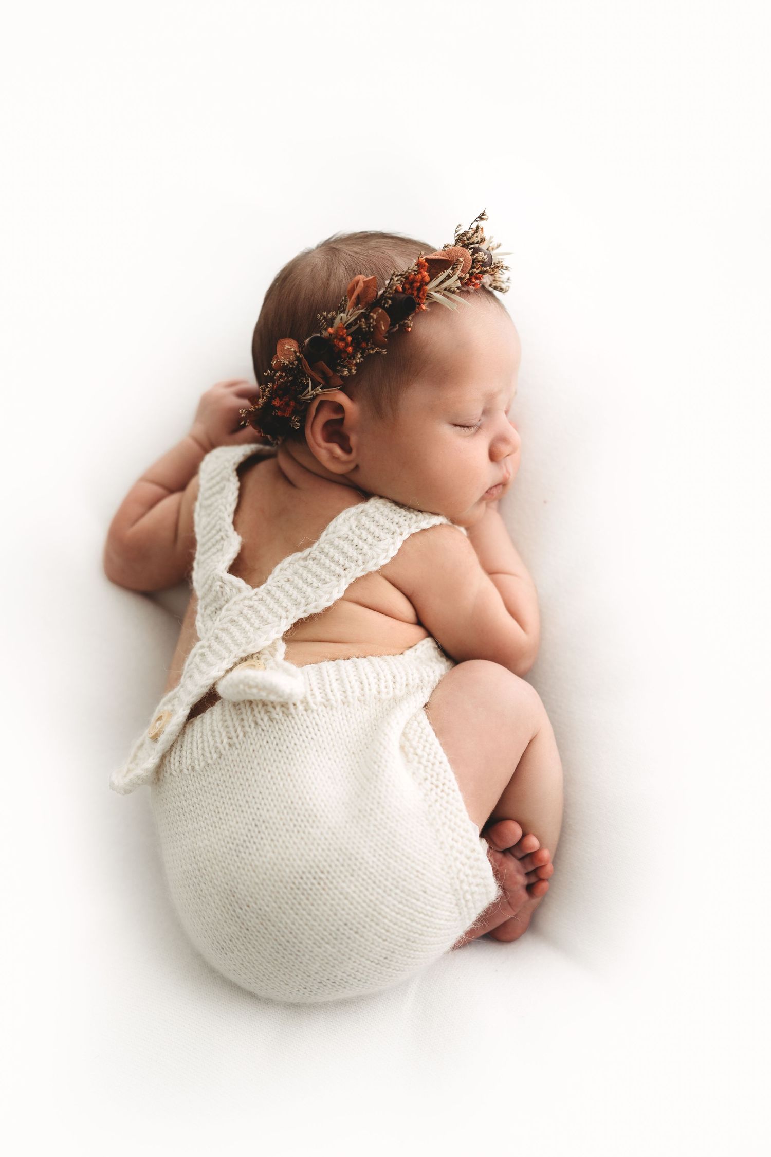 Sleeping newborn baby wearing floral crown and cream knit romper lying on white backdrop.