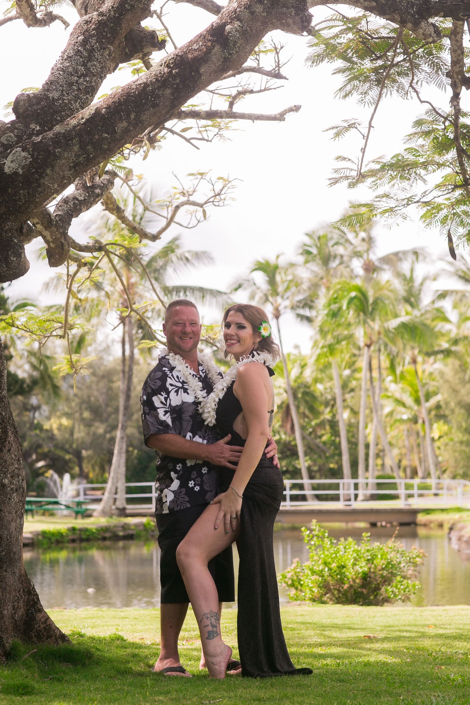 Smiling couple embracing under large tree in tropical Hawaiian garden with palm trees and pond.