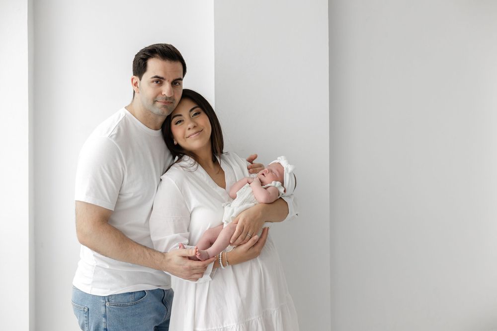Parents in white outfits share tender moments with their newborn baby during an intimate indoor photoshoot.
