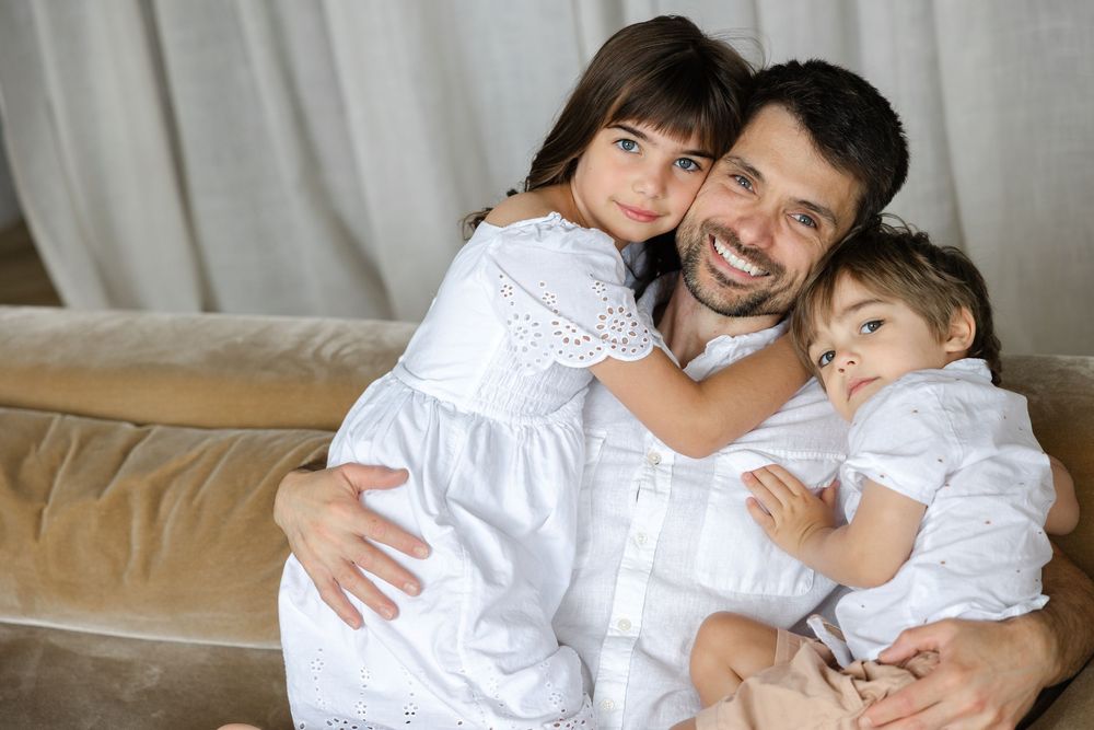 Family wearing coordinated white outfits cuddle and smile together while sitting on a beige couch.
