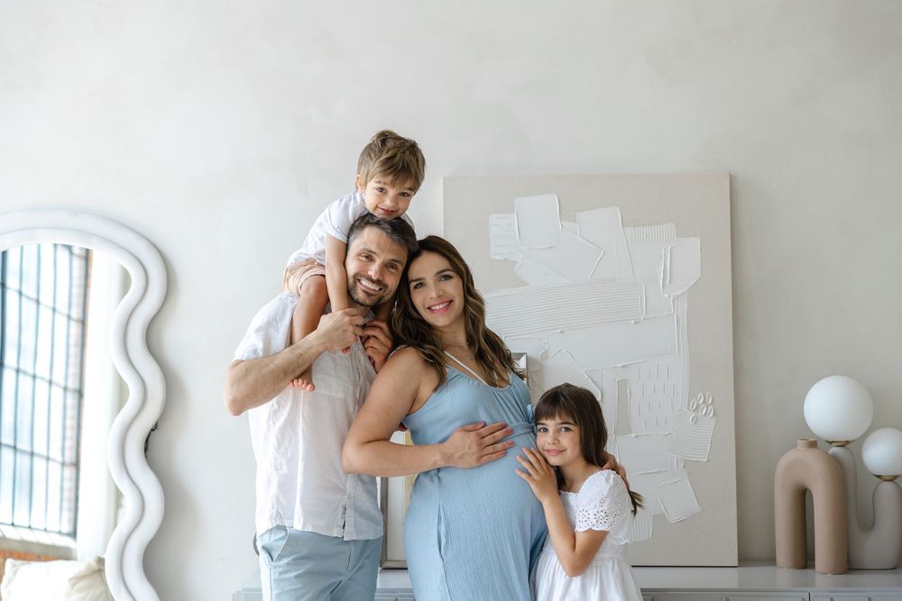Family wearing casual attire stands together smiling in front of white wall with decorative mirror and artwork.