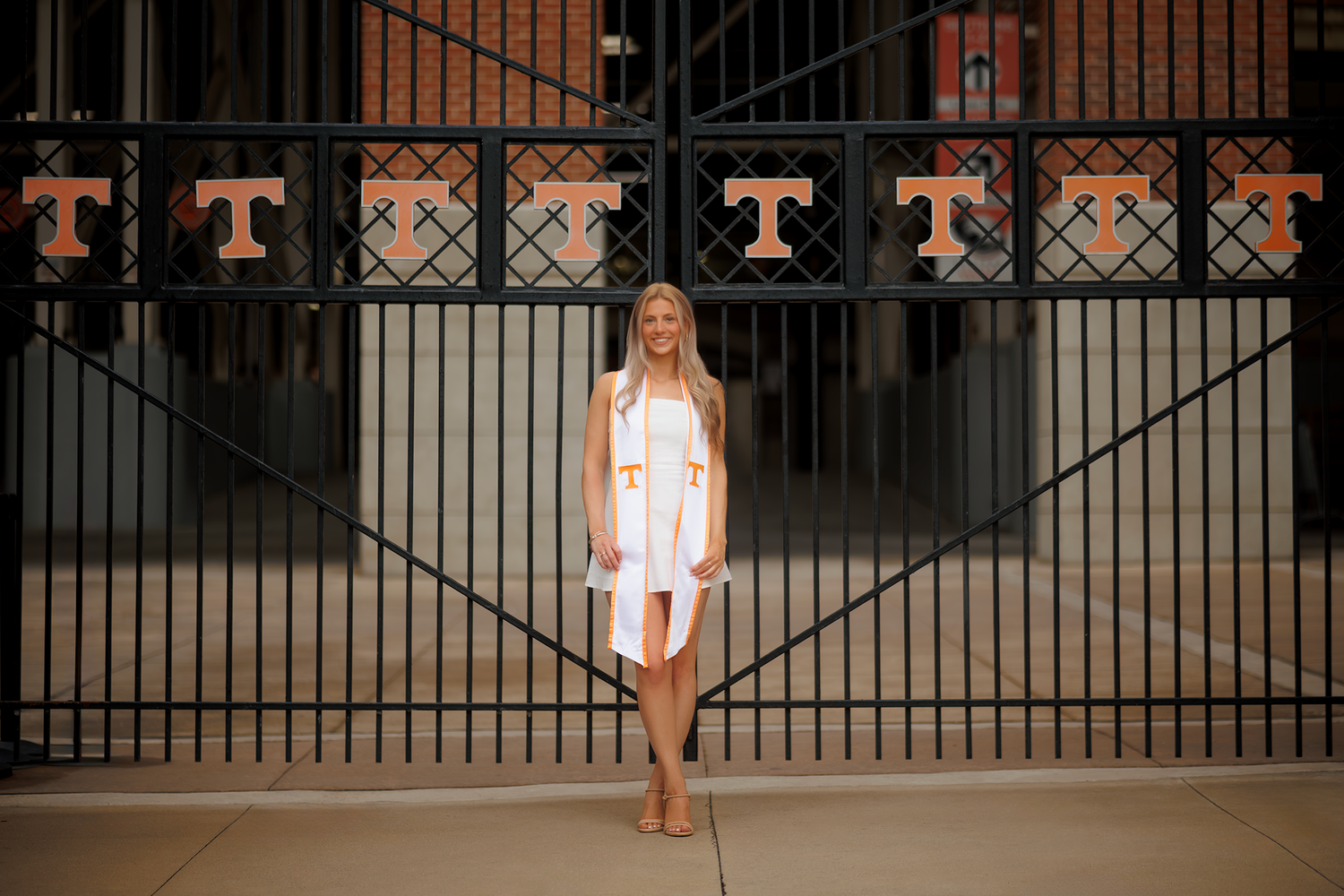 A person in a white dress stands in front of a large black iron gate with orange lettering at a stadium entrance.