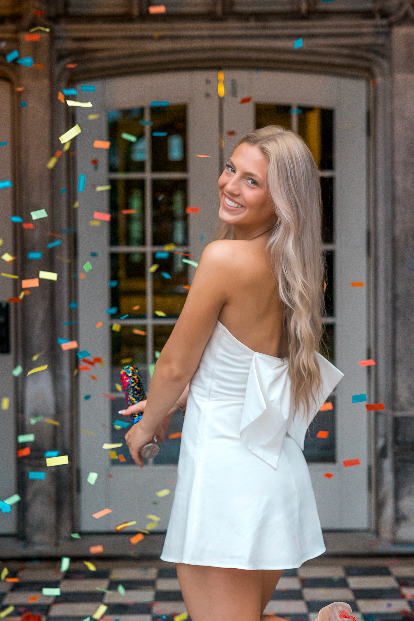 Someone in a short white strapless dress poses playfully as colorful confetti falls around them outside a building entrance.
