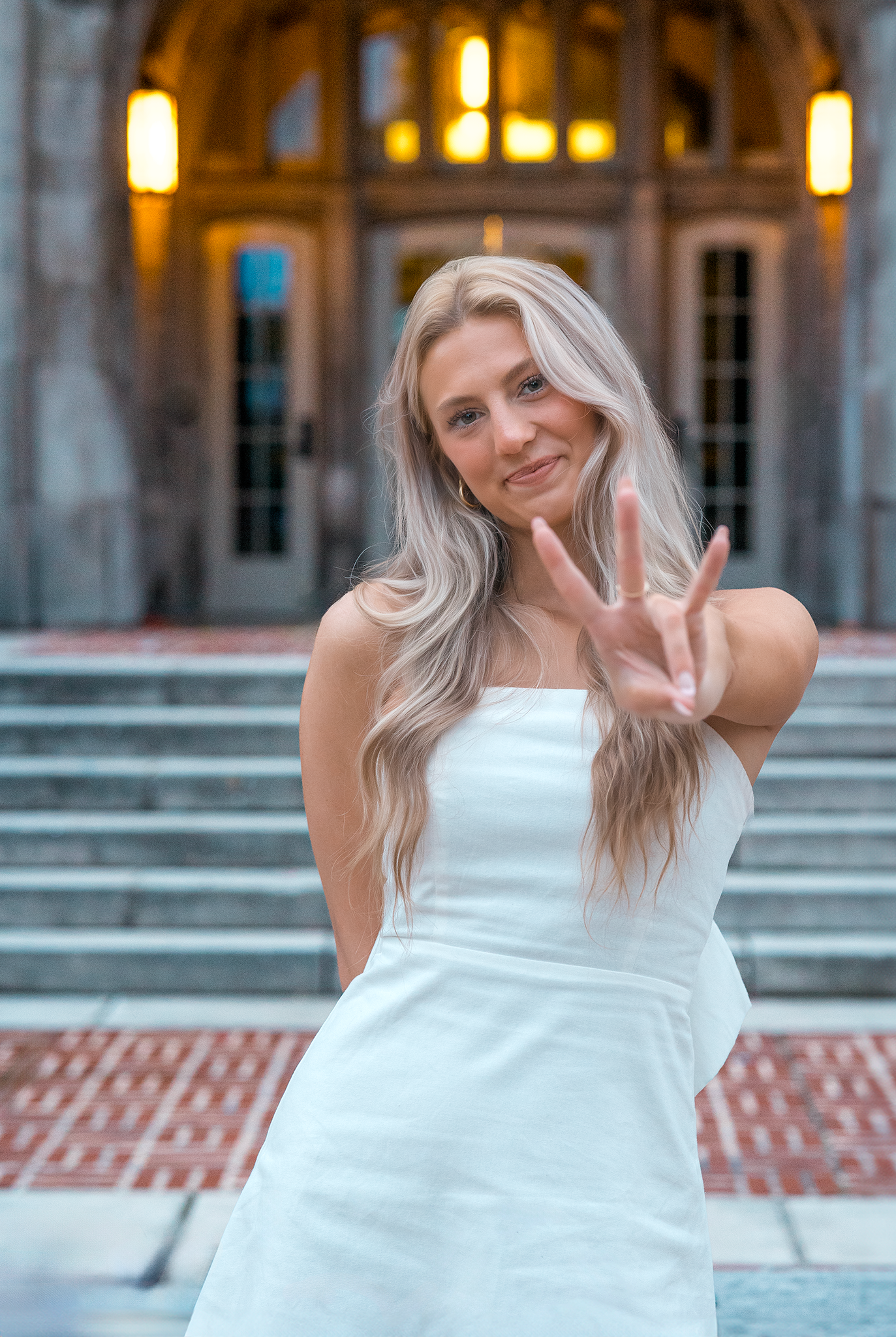 A person in a white summer dress makes a peace sign while standing on stone steps in front of an ornate building entrance.