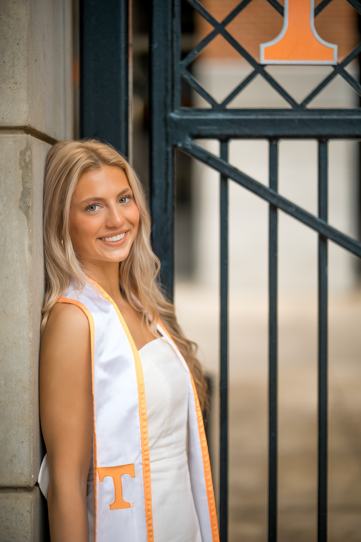 A graduate wearing a white dress with orange University of Tennessee graduation stole poses by an iron gate.