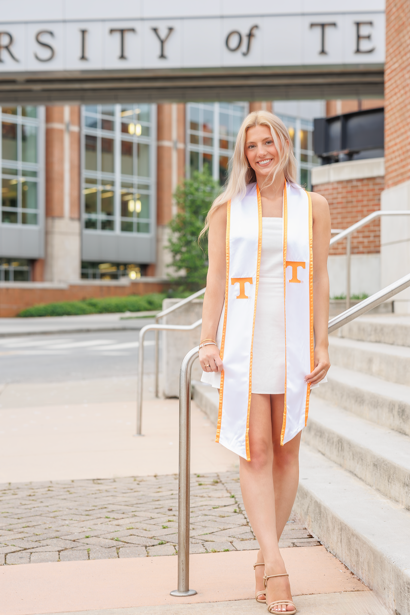 Graduate wearing white dress and orange University of Tennessee graduation stole stands on campus steps.