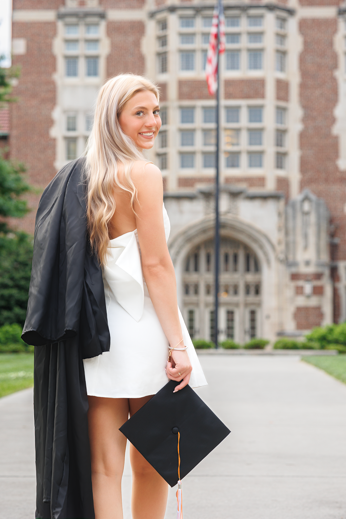 Graduate in white dress holding cap and gown stands in front of historic university building with American flag.