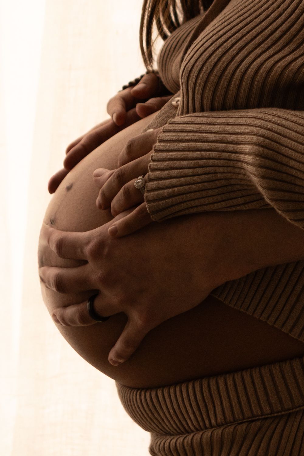 A young expecting mom is standing in front of a window, peering down at her belly. Her husband has placed his hands on her stomach with hers