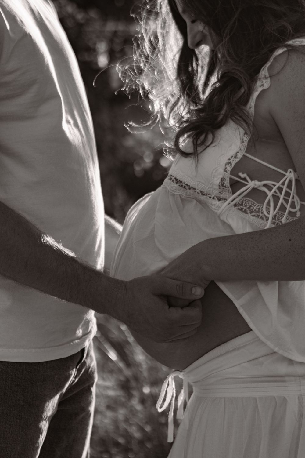 A young expecting couple wearing white looks down at mama's belly. Photographed in black and white.