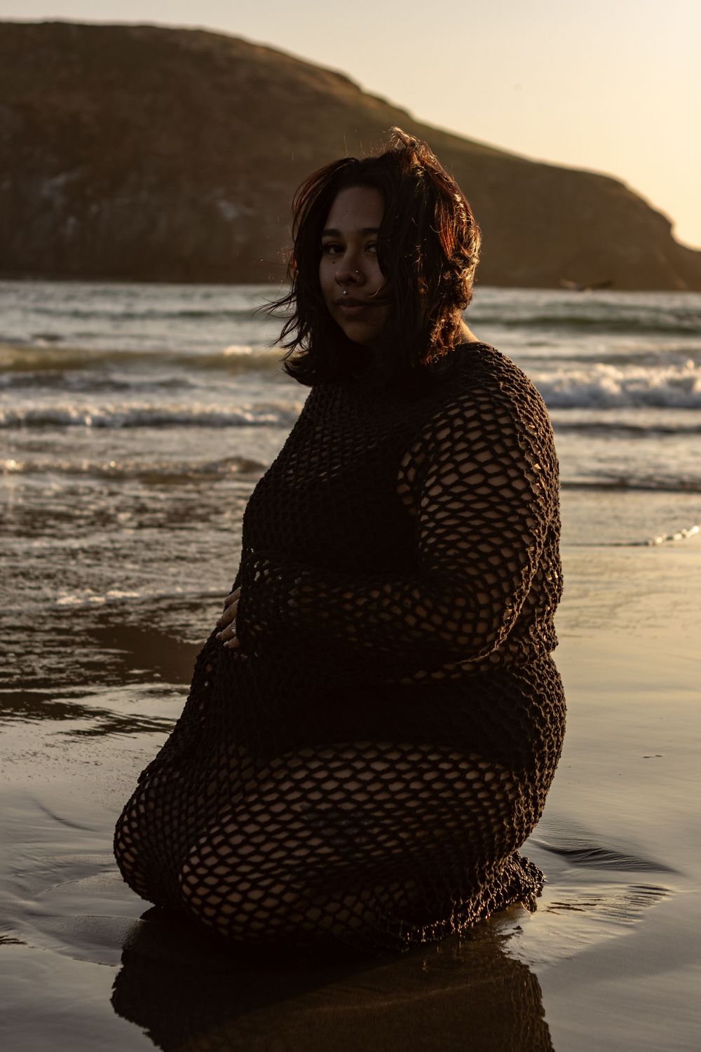 An expecting mama sits on the beach in all black, wearing a black two piece, and a long sleeve crochet coverup