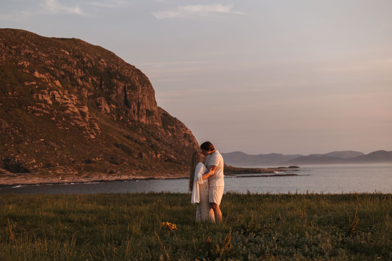 A couple stands together on a grassy cliff overlooking a serene coastal landscape at sunset.