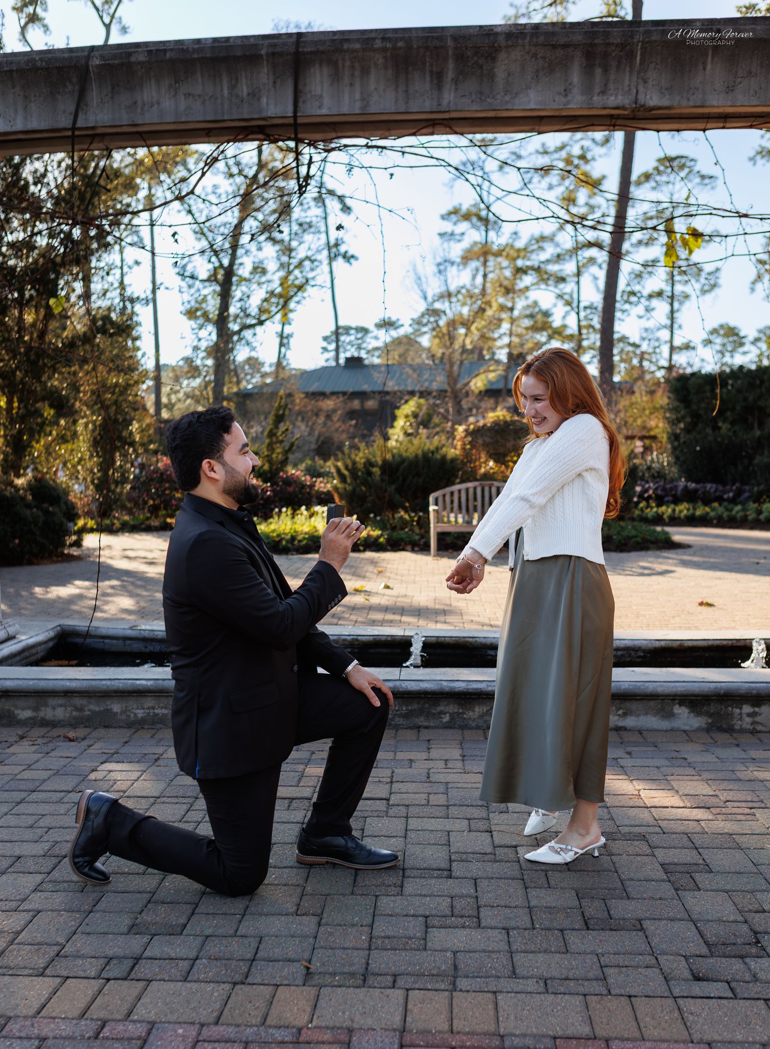 Man proposes on one knee to red-haired woman in white sweater at an outdoor garden plaza.