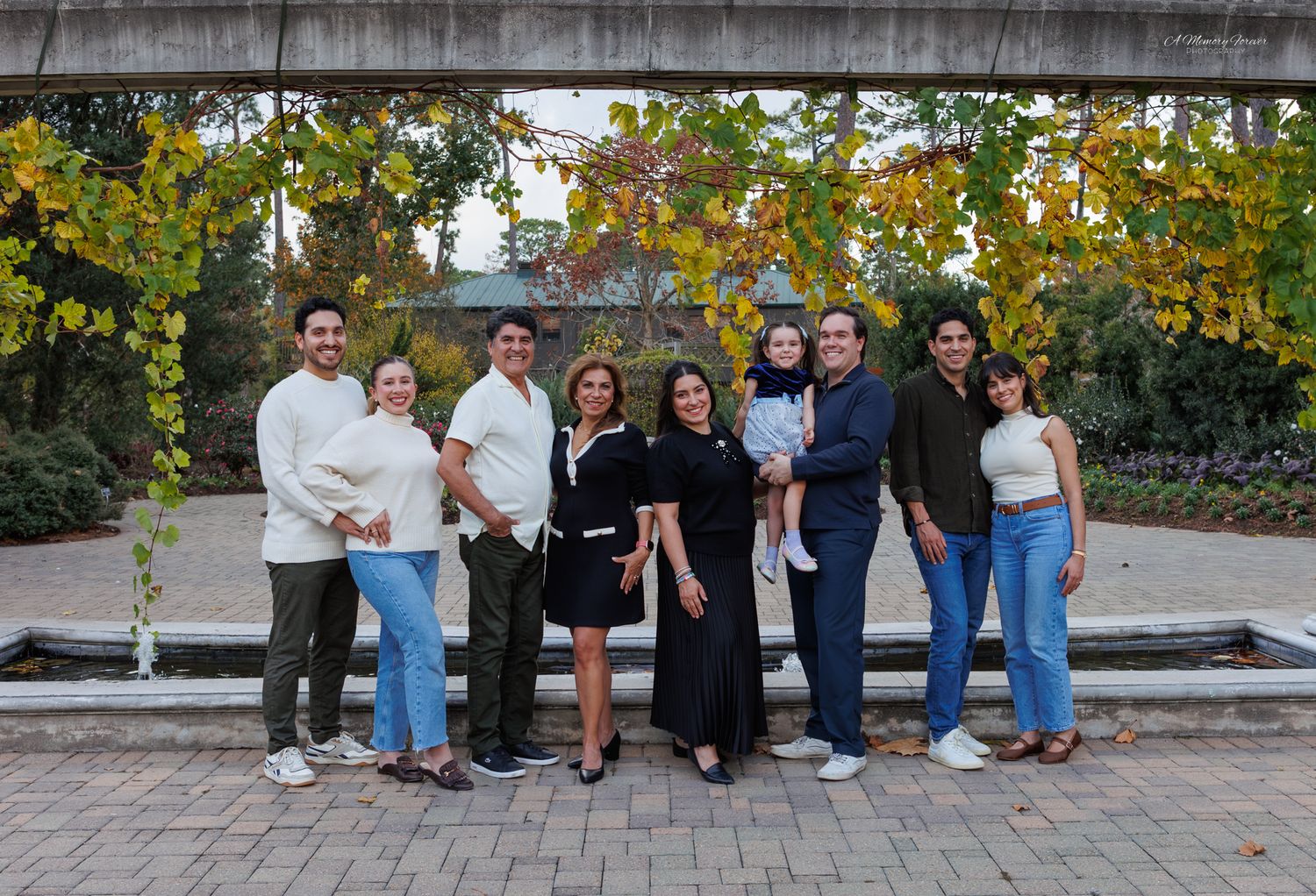 Large family group posing together outdoors near a fountain surrounded by autumn foliage in a park.