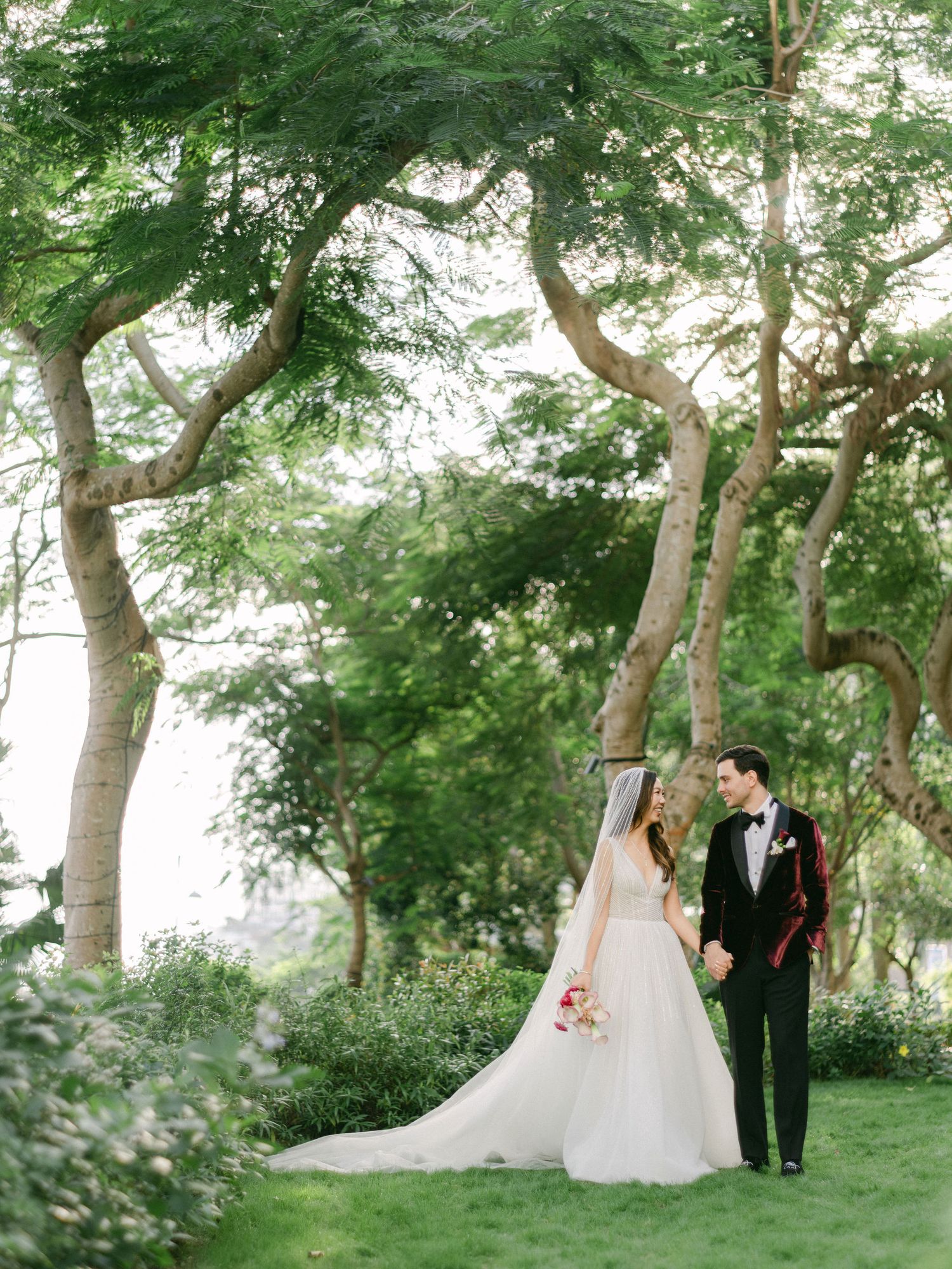 Bride and groom hold hands walking through lush green garden trees on their wedding day.