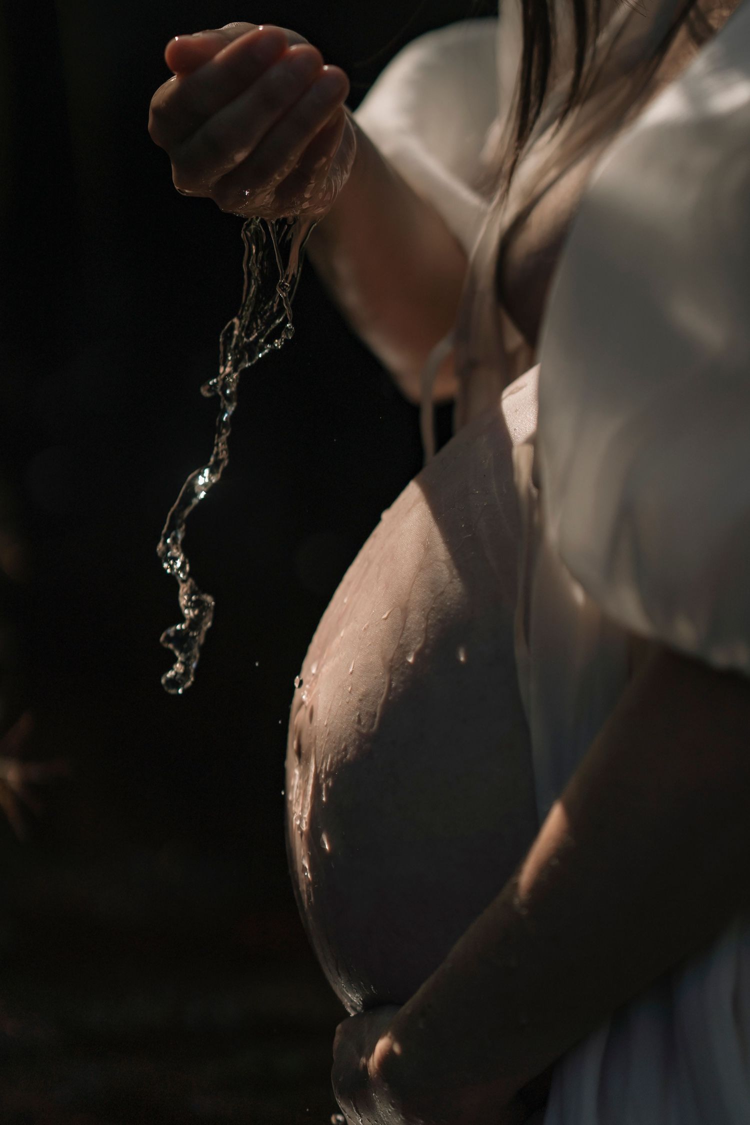 Pregnant woman in white dress letting water drip from her hand onto her bare baby bump in dramatic lighting.