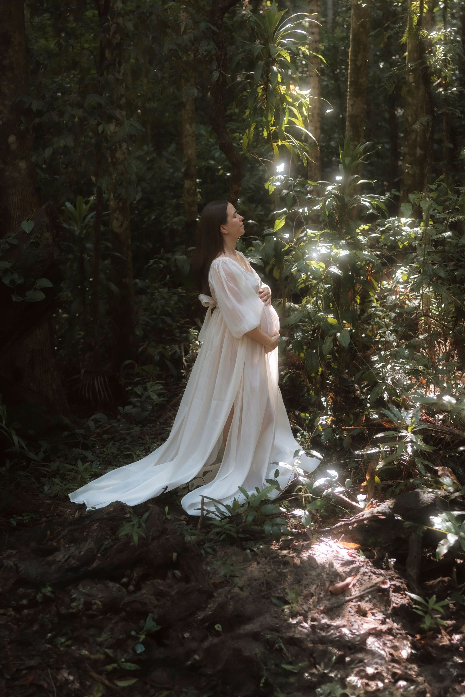 A pregnant woman in a flowing white dress stands in a sunlit forest setting, creating an ethereal maternity portrait.
