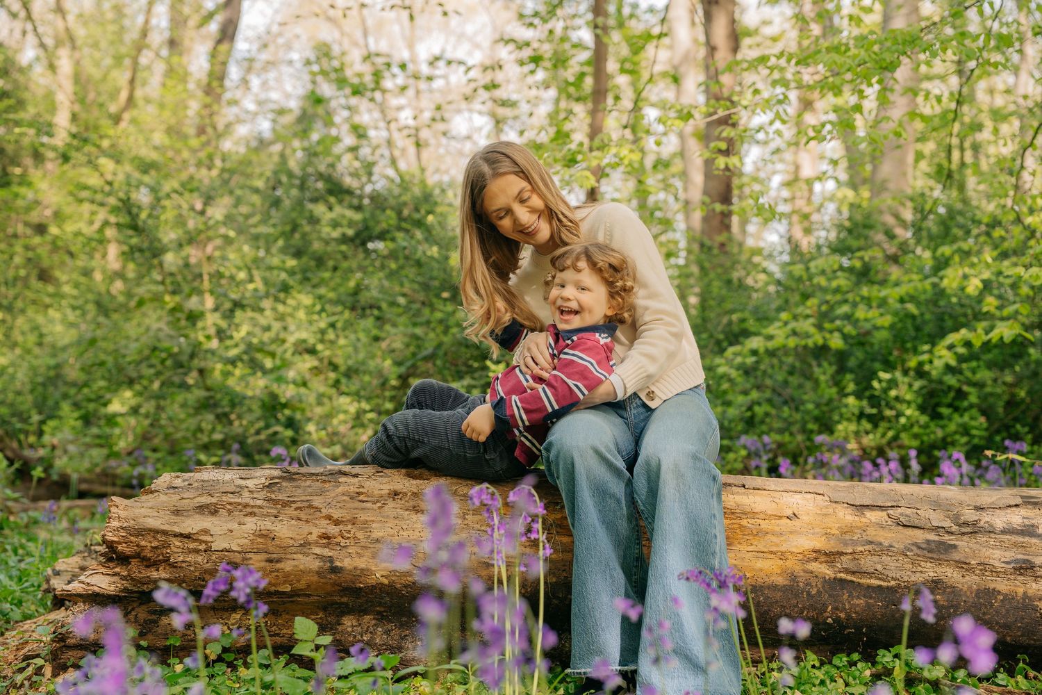 A mother and young child sit together on a fallen log surrounded by purple wildflowers in a lush green forest.