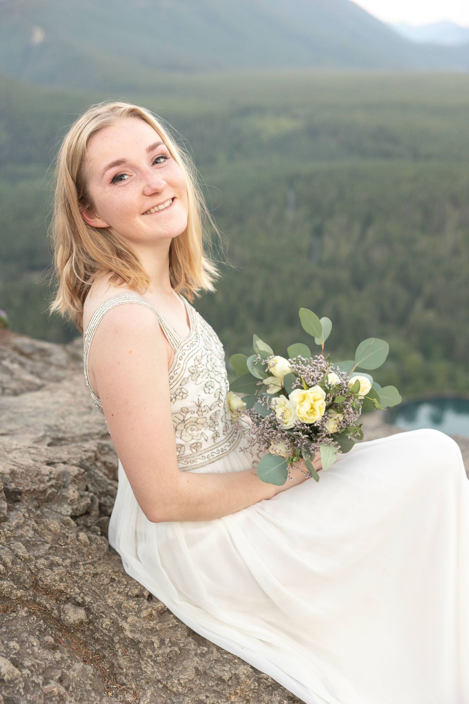 Bride in sequined wedding dress sits on mountain cliff holding rustic bouquet with yellow roses and eucalyptus.
