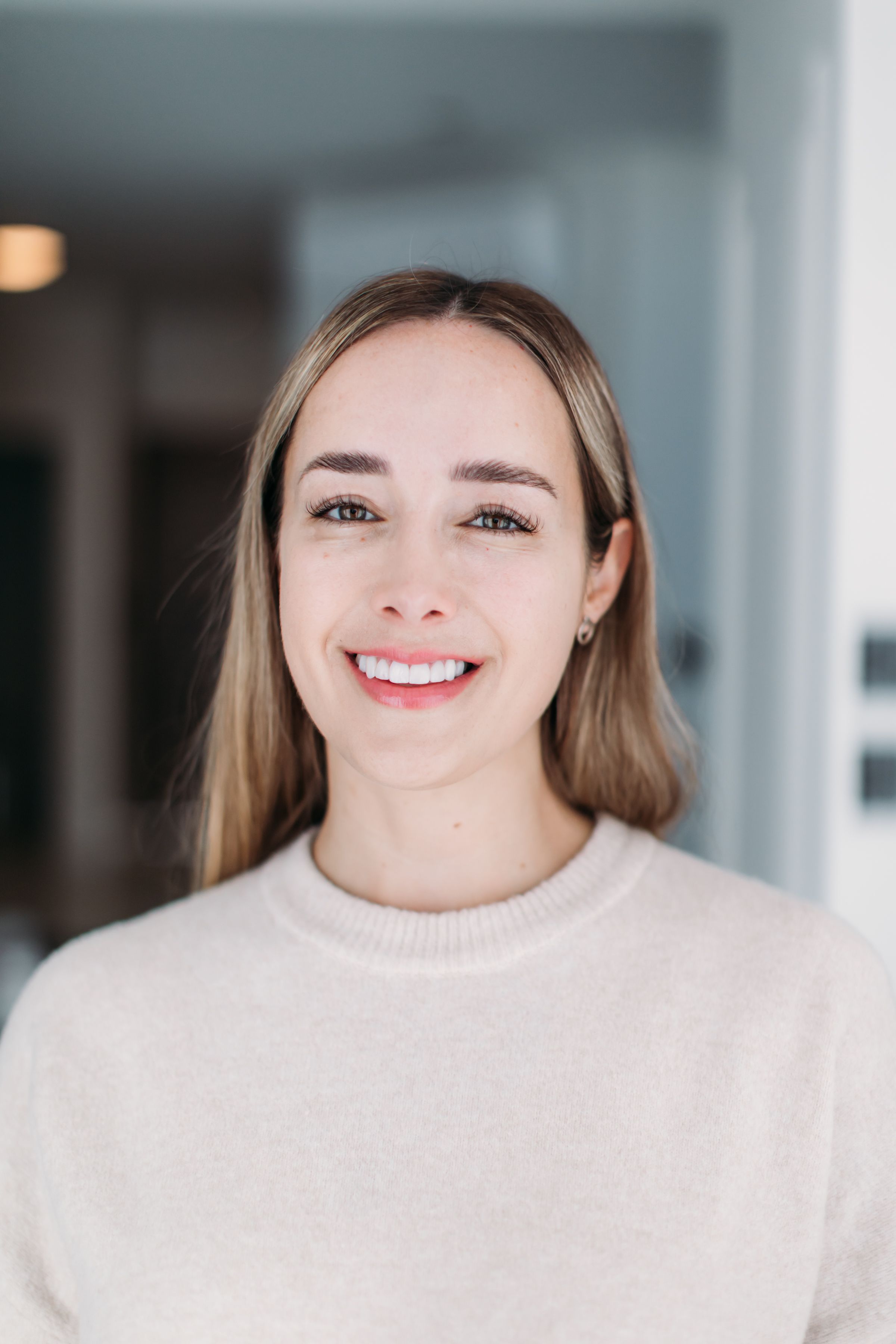 Professional headshot of a smiling person wearing a cream sweater against a bright interior background.