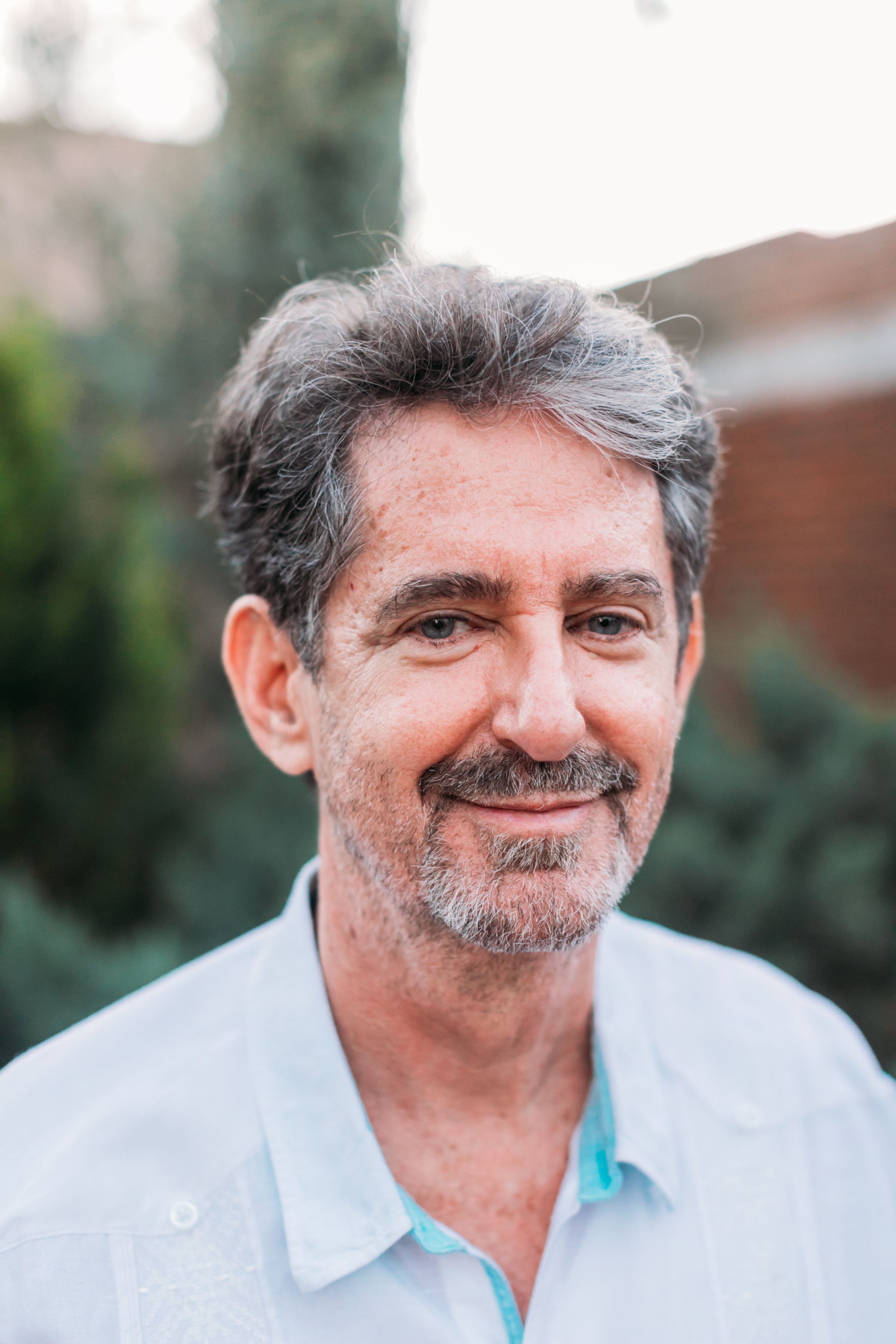 Close-up outdoor portrait of a person with grey hair and beard wearing a light blue shirt.