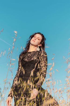 Fashion portrait in black lace dress among tall grass against bright blue sky.