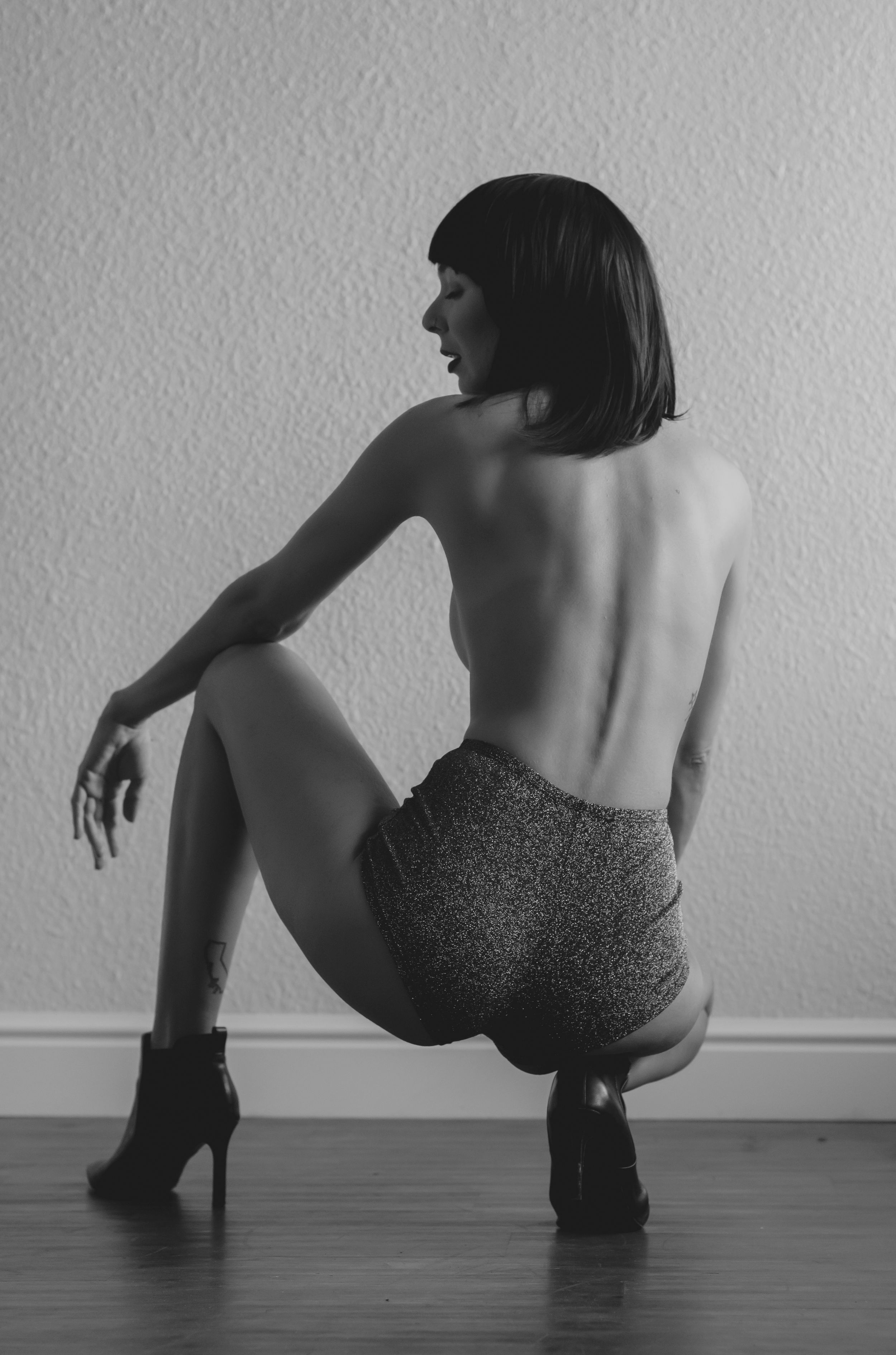 Black and white artistic photo of a figure in shorts and heels posing on a wooden floor.