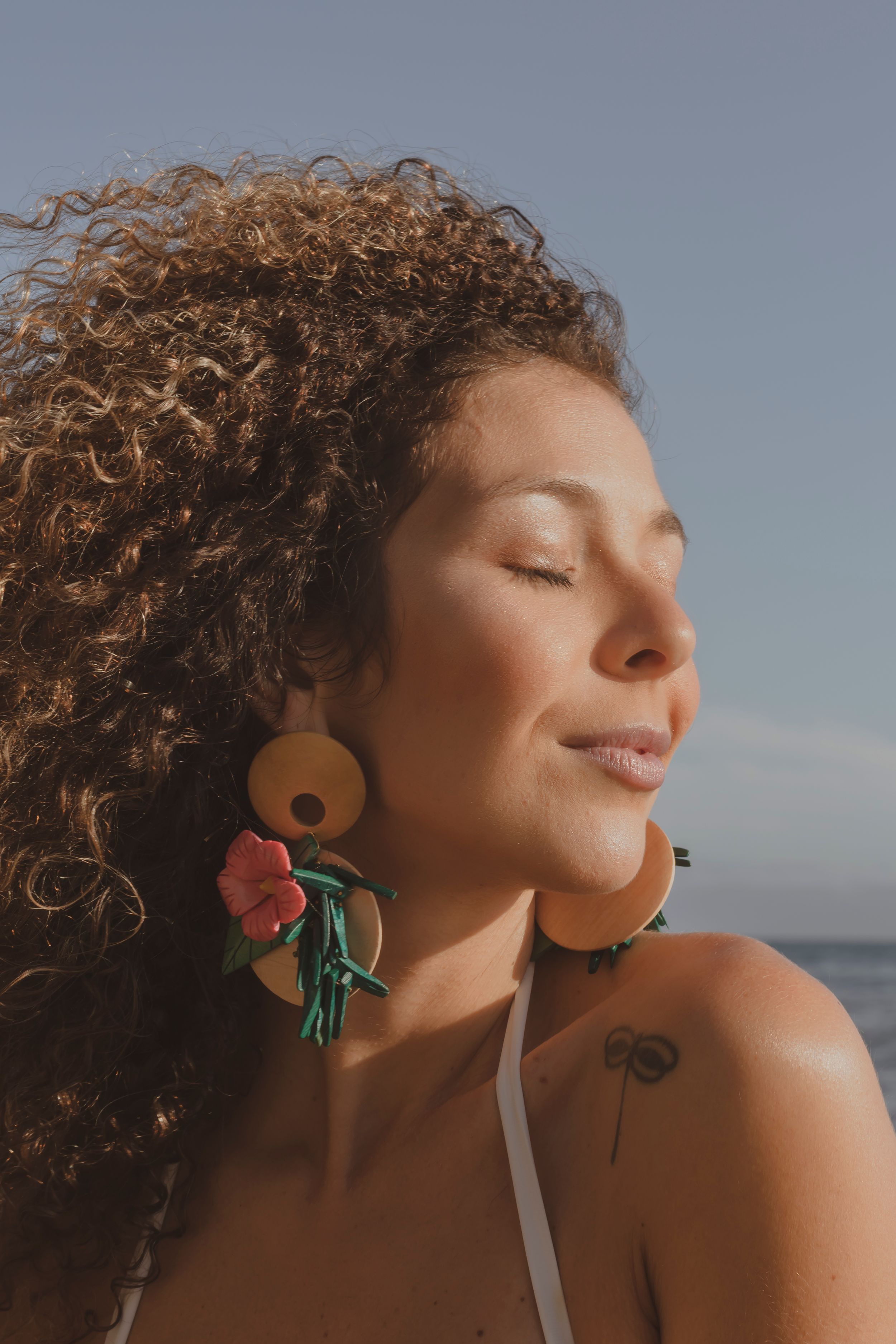 Sun-kissed portrait featuring curly hair and colorful floral earrings against an ocean backdrop.