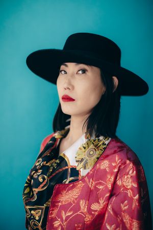 Portrait against turquoise background featuring traditional Asian attire and wide-brimmed black hat.