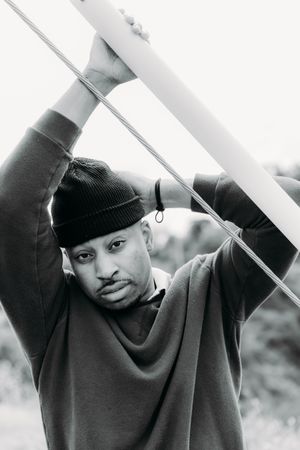 Black and white portrait of a person in a beanie and sweater leaning against railings outdoors.