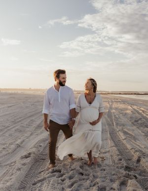 A couple in casual attire walks together on a sandy beach at sunset, with the woman wearing a flowing white dress.