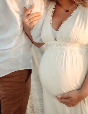A close-up photo of a white maternity dress with delicate lace trim and pearls, showing a pregnant belly being cradled.