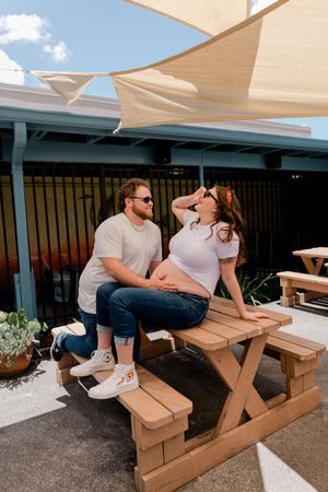 Pregnant couple sitting on wooden picnic table under shade sails in sunny outdoor patio area.
