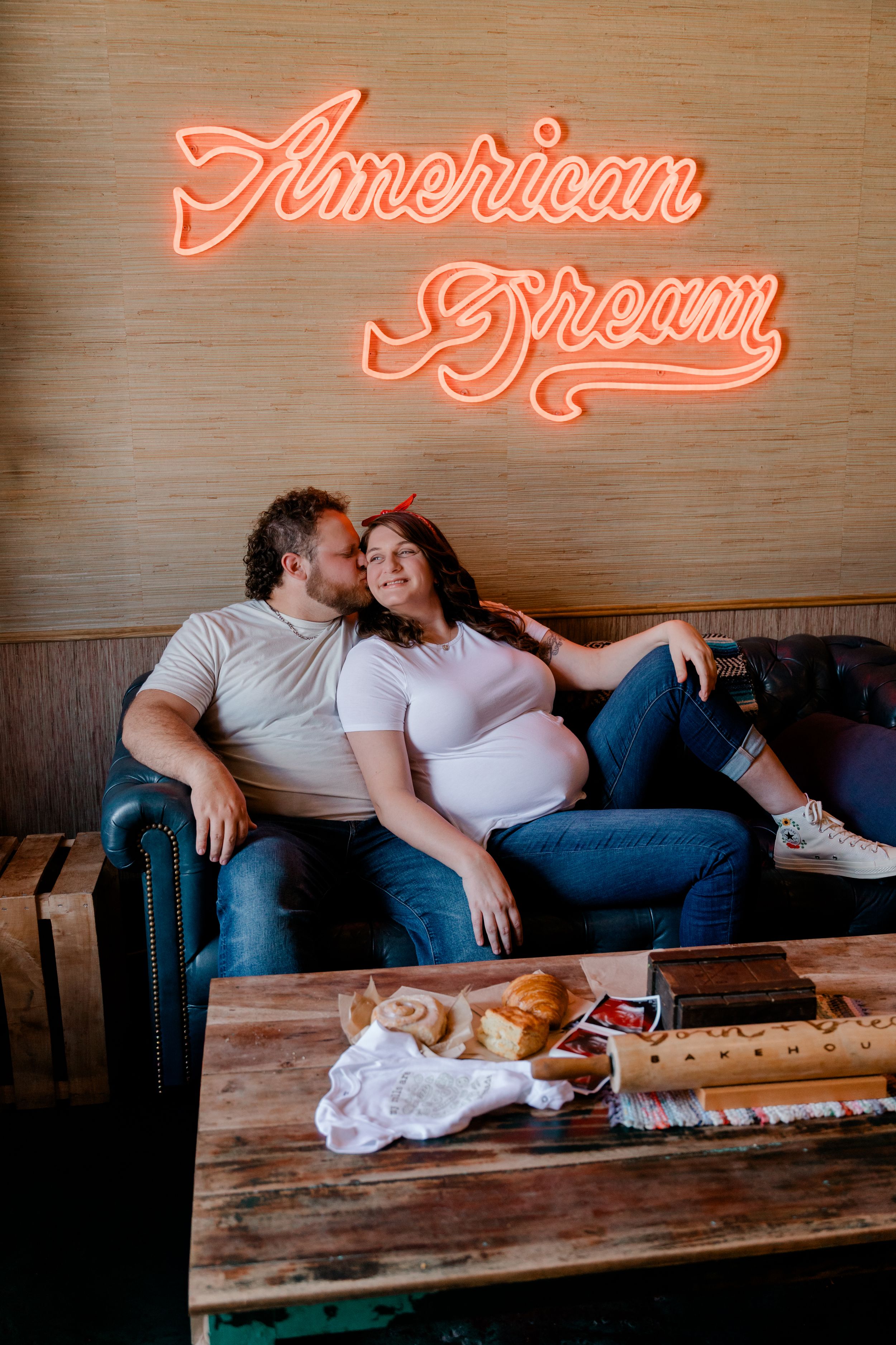Pregnant couple sitting on leather sofa under American Dream neon sign with bread on wooden table.