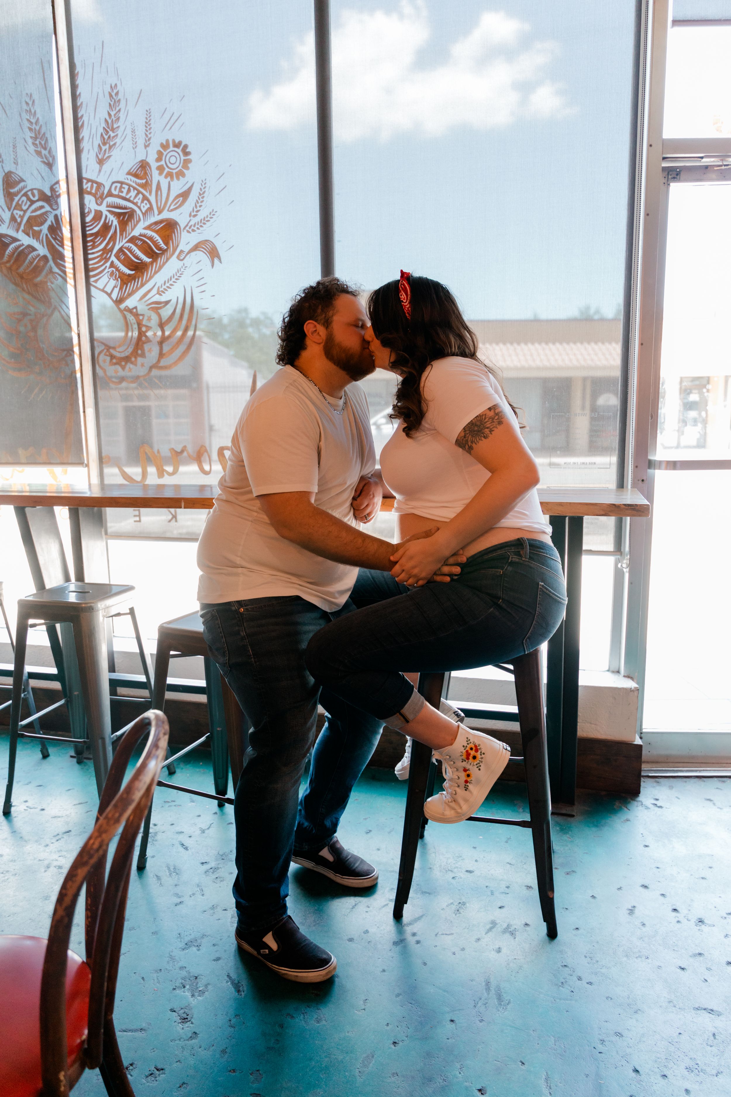 Couple sharing a kiss on bar stools inside a cafe, both wearing white shirts and jeans.
