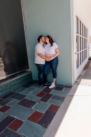 Couple sharing a kiss against a mint green wall on a colorful tiled sidewalk, woman showing a baby bump.