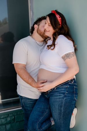 Pregnant couple in white tees and jeans share a tender moment, woman with red bandana headband showing baby bump.
