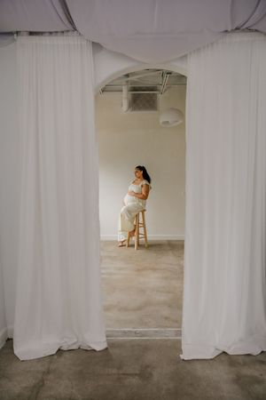 A figure in white sits on a wooden stool, framed by flowing white curtains in a minimalist studio space.