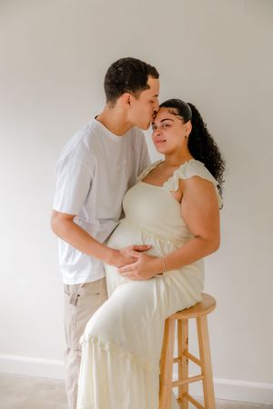 A couple in white outfits share an intimate moment in a minimalist setting with soft natural lighting.