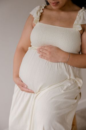 A pregnant person in a white smocked dress gently cradles their belly while wearing a delicate bracelet.