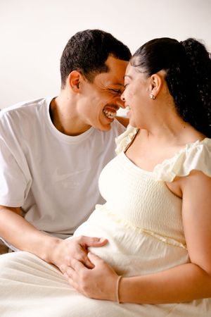 A loving couple laughs intimately together while sitting in casual white clothing against a light background.