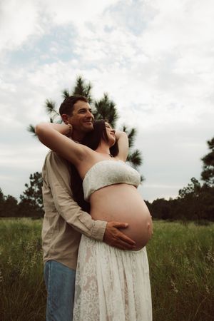 Romantic maternity photoshoot in a natural field setting featuring a white lace dress and tender embraces.