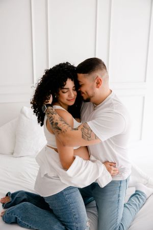 Intimate couple embraces on white bedding wearing jeans and casual attire while showing their tattoos.