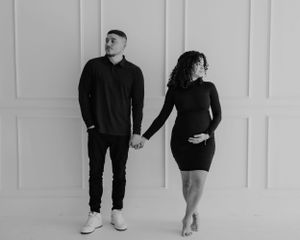 A black and white photo of a couple holding hands against a white wall, with both wearing dark outfits.