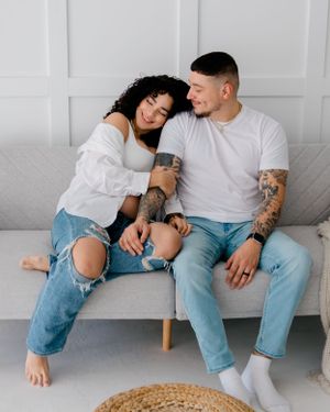A couple in matching white tops and ripped jeans cuddle on a gray couch while sharing an intimate moment at home.