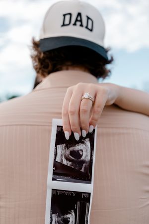 Pregnant couple pregnancy announcement with ultrasound photos, man wearing DAD hat, woman showing engagement ring.