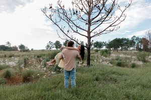 Couple embracing near a bare tree in a green meadow under a cloudy sky.